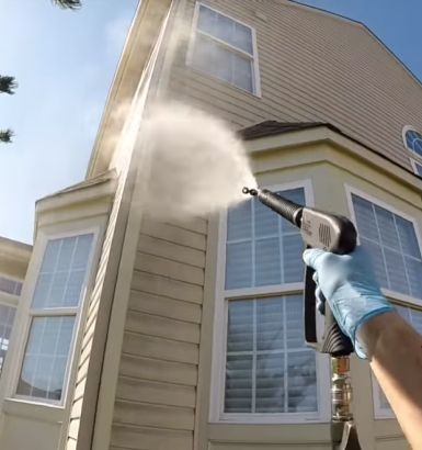Person wearing gloves pressure washing the siding of a house with a high-pressure water jet.